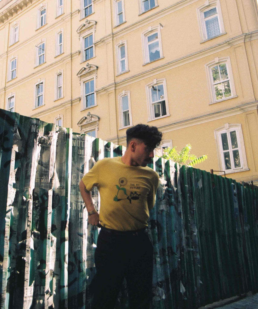 a man is wearing Aponia big picture yellow tshirt and standing in an alley while sun shines from one side  