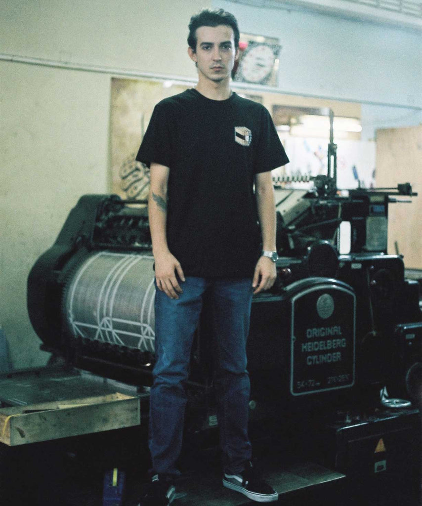 A boy is wearing Aponia Book Bomb black t-shirt and standing in front of a printing press 