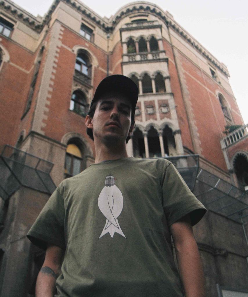 A guy is standing in street with a high classic building in back and wearing an olive t-shirt with graphic of a bird with a cage on head.
