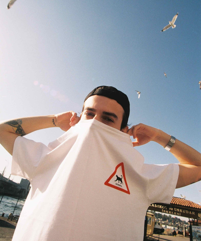A guy is masking half of his face with his worn Aponia white cat t-shirt with graphic of a walking cat in triangle traffic sign and underwritten of Istanbul
