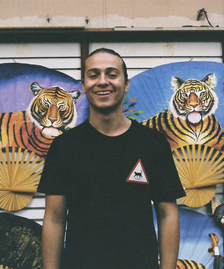 A smiling guy is standing in front of a wall with painting of tigers and wearing a black Istanbul Cat t-shirt of Aponia Store.