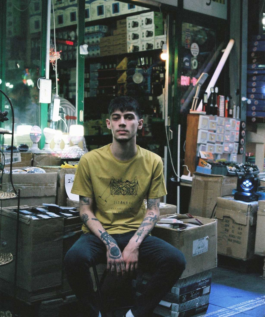 A guy is wearing Aponia cat's cradle yellow t-shirt and sitting in front of a store in bazaar 